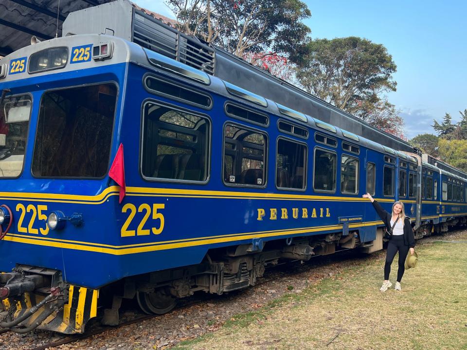 Lindsay poses with an arm raised in front of a blue PeruRail train with yellow accents.