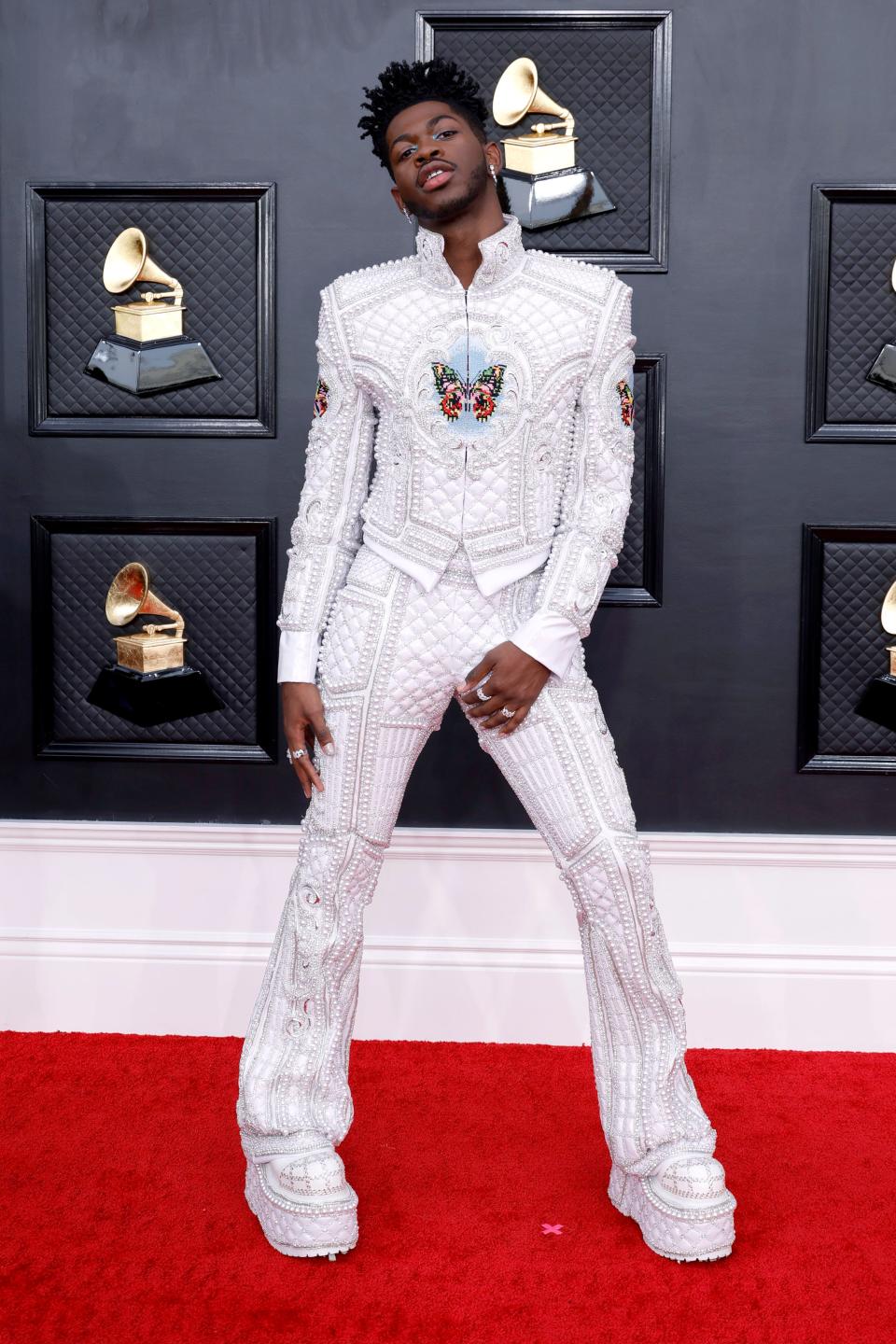 Lil Nas X in a white textured and architectural 3D jacket and pants with butterfly images on the chest and arms. He's wearing white matching platform boots.