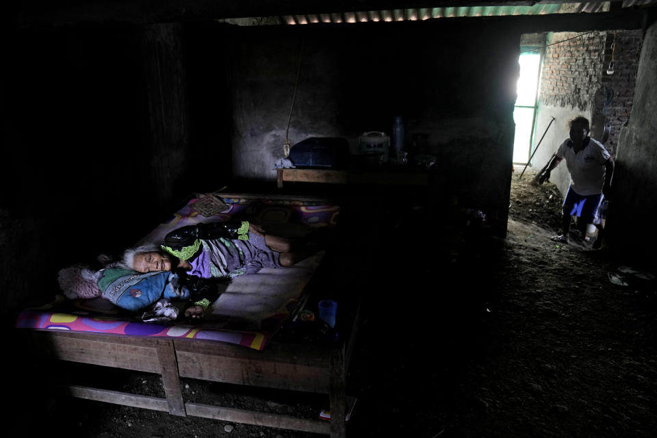 Suratmi, who is paralyzed, lies in her bed as her daughter Ngatiroh, right, walks on the floor that has been raised to prevent flood water in their house in Timbulsloko, Central Java, Indonesia, Saturday, July 30, 2022. Ngatiroh said that after her mother Suratmi's home collapsed due to flooding, she moved her into the windowless, dirt-floor home that they now share. (AP Photo/Dita Alangkara)
