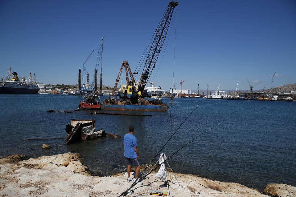 A man fishes as a floating crane prepares to raise a shipwreck, on Salamina island, west of Athens, on Monday, Aug. 26, 2019. Greece this year is commemorating one of the greatest naval battles in ancient history at Salamis, where the invading Persian navy suffered a heavy defeat 2,500 years ago. But before the celebrations can start in earnest, authorities and private donors are leaning into a massive decluttering operation. They are clearing the coastline of dozens of sunken and partially sunken cargo ships, sailboats and other abandoned vessels. (AP Photo/Thanassis Stavrakis)