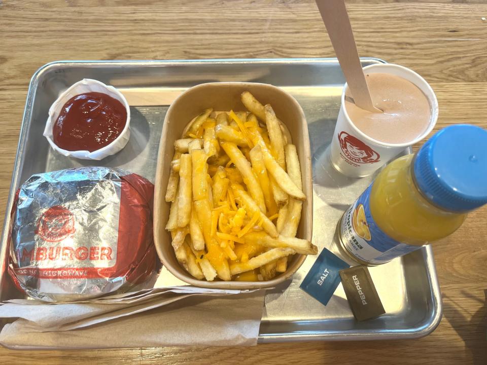 A tub of ketchup, a burger, cheesy chips, a brown slush, a bottle of juice.