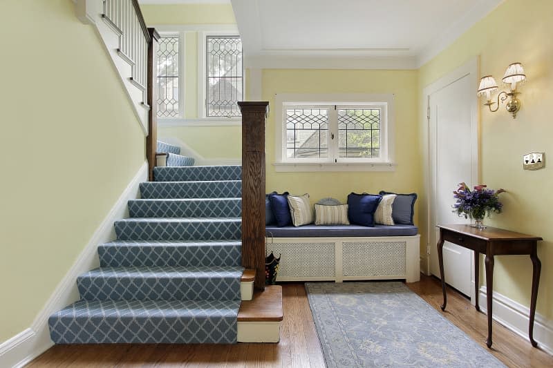 Bench with blue cushion and throw pillows next to carpeted stairway in entryway with yellow walls. 62935273