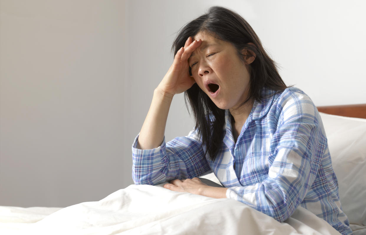 A woman sits up in bed, propping herself up on her arm, her eyes closed, yawning.