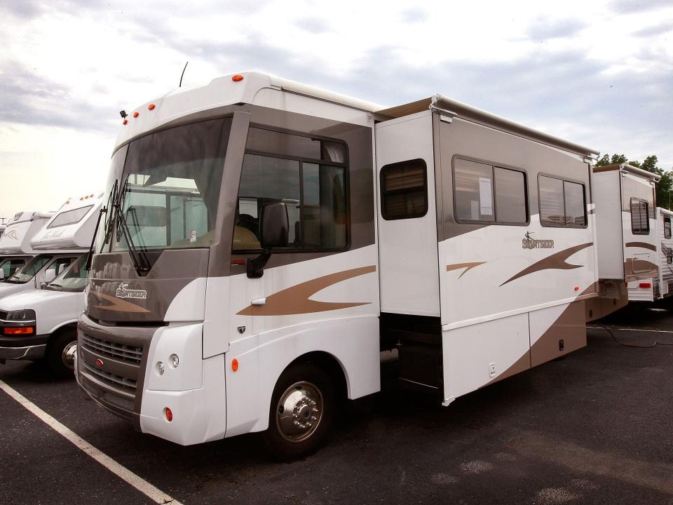 A large RV is parked in a lot with other RVs behind it and cloudy skies.