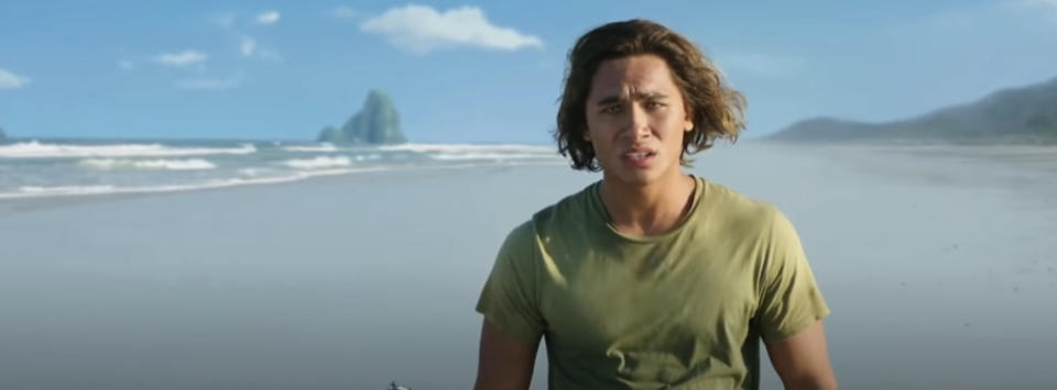 Young man with wavy hair stands on a beach looking distressed, wearing a green T-shirt. The scene is from a movie or TV show