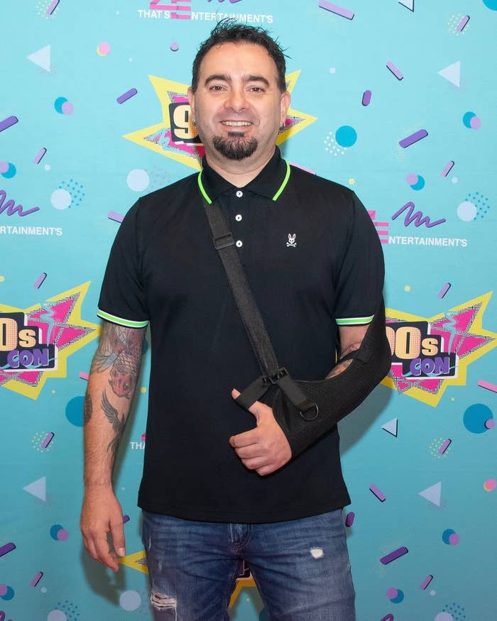 Chris Kirkpatrick smiles on the red carpet of 90s Con. His left arm is in a slin