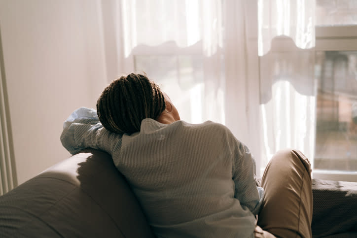 Person with braided hair sitting on a couch, looking out a window with sheer curtains, with their arm resting on the couch back