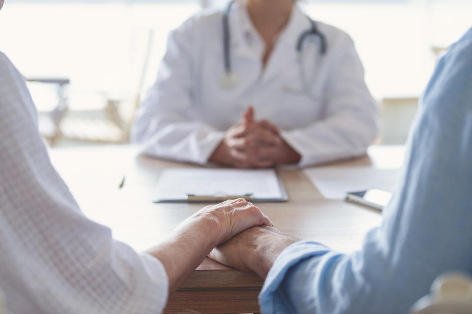 Mature couple holding hands at a doctors office. Doctor can be seen in the background.