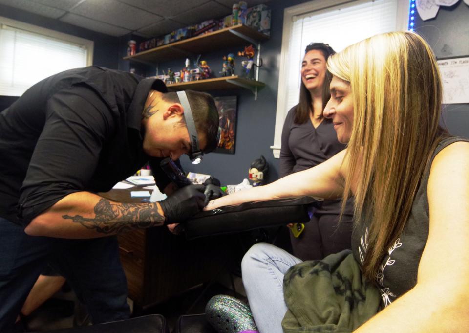 Manuel Barros of Plymouth, tattoo artist at K&M Studio in Brockton, works on the arm of client Angie Clem, right, of Weymouth, as friend Megan Berube of Braintree looks on.