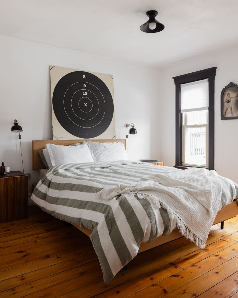 Target art above bed with striped bedding in white bedroom with wood flooring.