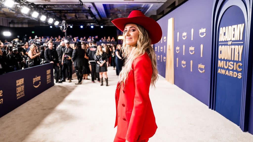 lainey wilson smiles at the camera while looking over one shoulder, she stands on a tan carpet in an all red outfit as photographers and other people stand in the background