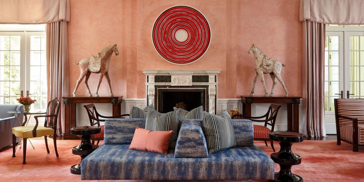 a pink walled and rugged living room with large red circular art over  white marble fireplace mantel with nothing on it and two tall wooden white saddled horses on three foot dark tables flanking the fireplace and facing it  as tall simple cream valanced curtain over french doors right next to each and in the center of the room is a large lounge sofa with blue stripped fabric and heaped with pillows