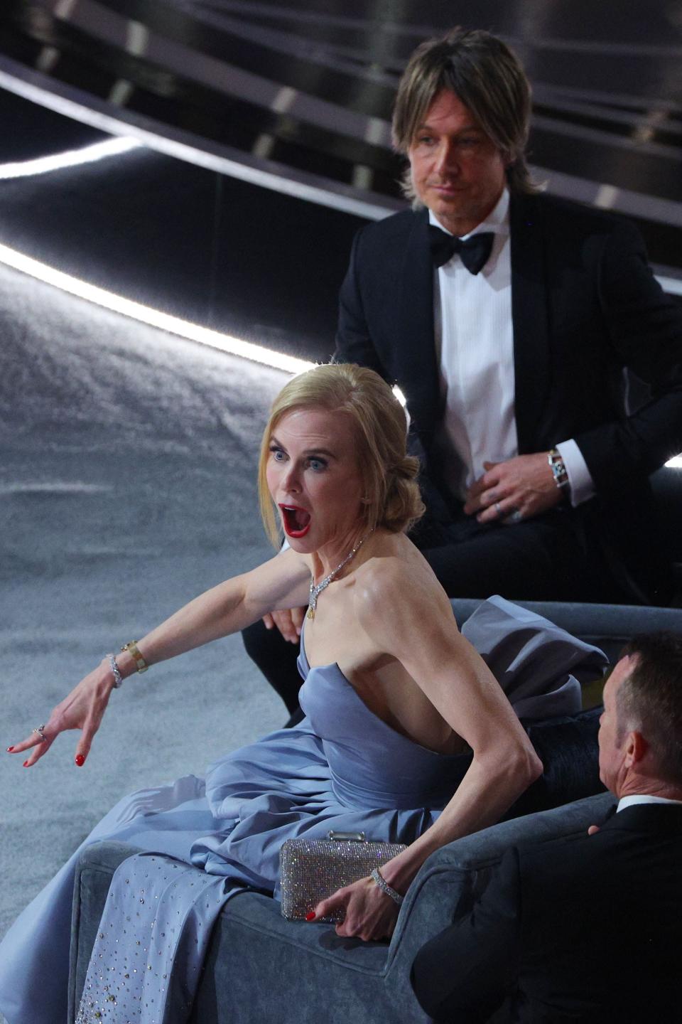 a photo of nicole kidman expressively reacting to something, twisted slightly in a chair with her eyes wide and eyebrows raised and mouth open. she's at the oscars wearing a blue gown, and her husband keith urban is behind her