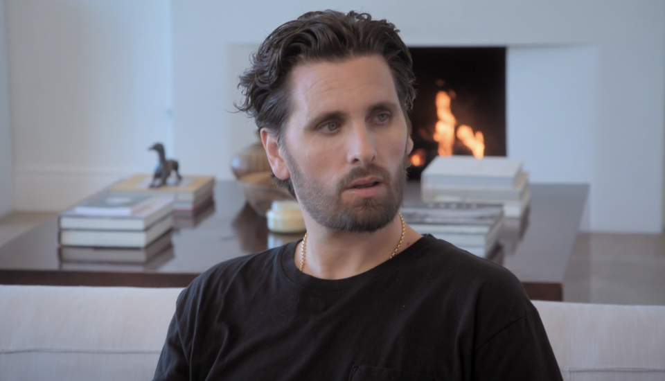 Scott Disick sits indoors with a pensive expression, wearing a casual black T-shirt. A fireplace and stacked books are visible in the background