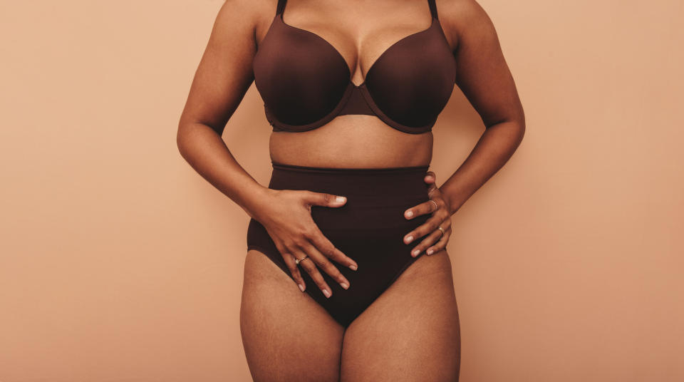 A person stands against a neutral background, wearing underwear and a bra. Their hands are placed on their hips
