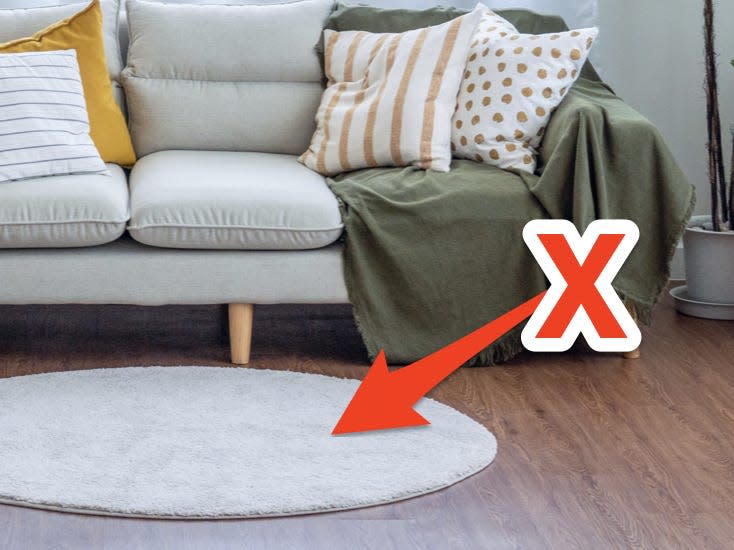 Gray couch with green throw beside houseplants with white, basic rug on wooden floor and red X and arrow pointing to rug