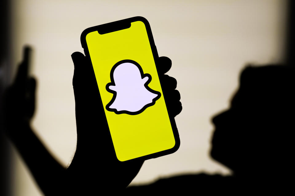 Snapchat logo displayed on a cellphone screen, with the silhouette of someone holding a cellphone. 