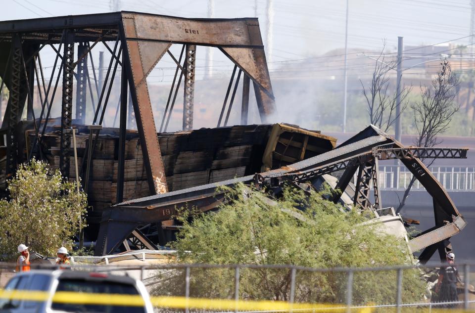 A derailed freight train burns on a bridge spanning Tempe Town Lake as part of the collapsed bridge bends in the foreground Wednesday, July 29, 2020, in Tempe, Ariz. Officials say a freight train traveling on the bridge that spans a lake in the Phoenix suburb has derailed, setting the bridge ablaze and partially collapsing the structure. (AP Photo/Ross D. Franklin)