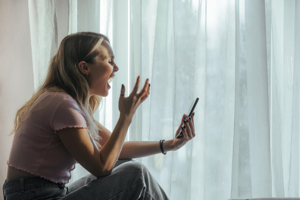 Woman sitting by window, excitedly gesturing at her phone. She's in a casual outfit