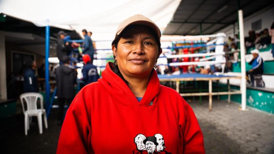 A woman in a red jumper and beige cap looks at the camera