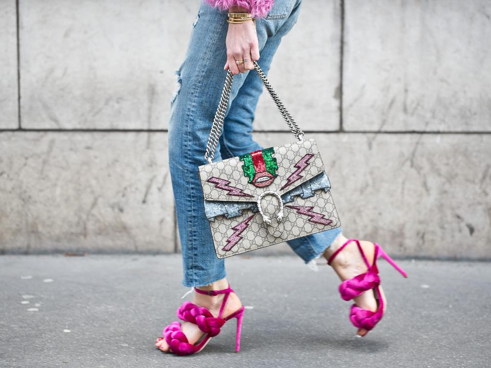 person walking down the street wearing blue jeans and puffy pink high heels while carrying a sparkly purse with lots of patches