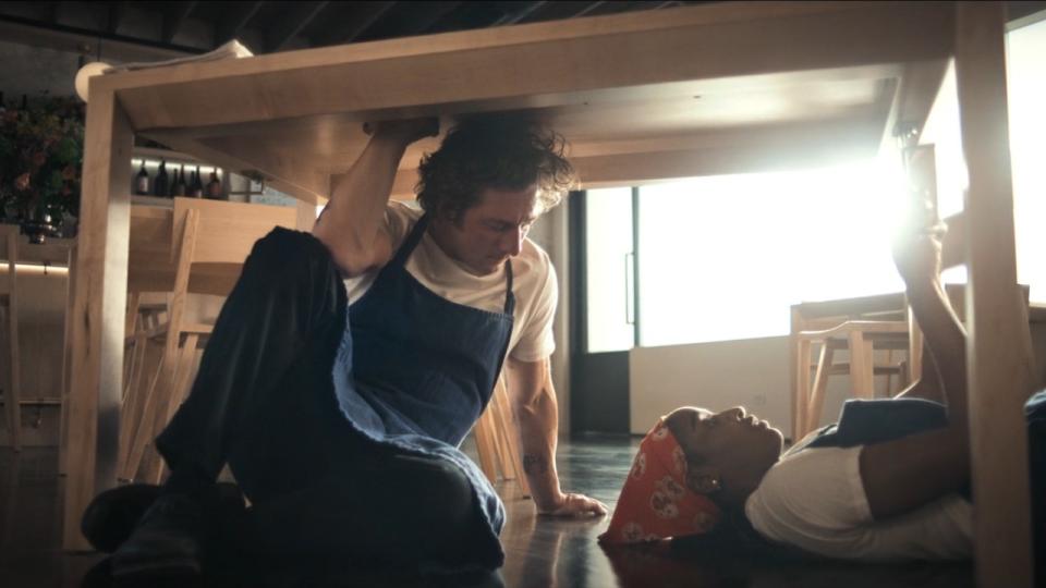 Jeremy Allen White sitting under a table and Ayo Edebiri laying under the table.