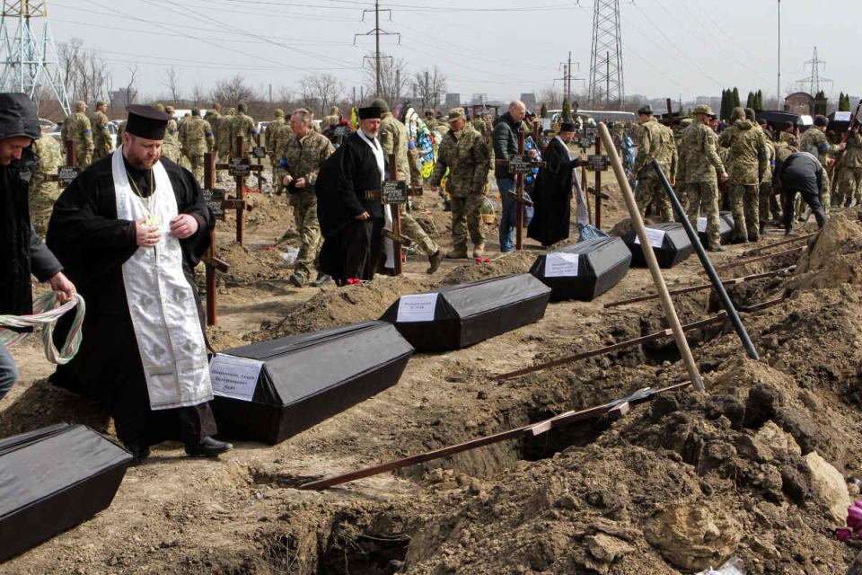 Priester und Soldaten stehen auf einem Friedhof in Dnipro neben Särgen vor ausgehobenen Gräbern während einer Beisetzung.