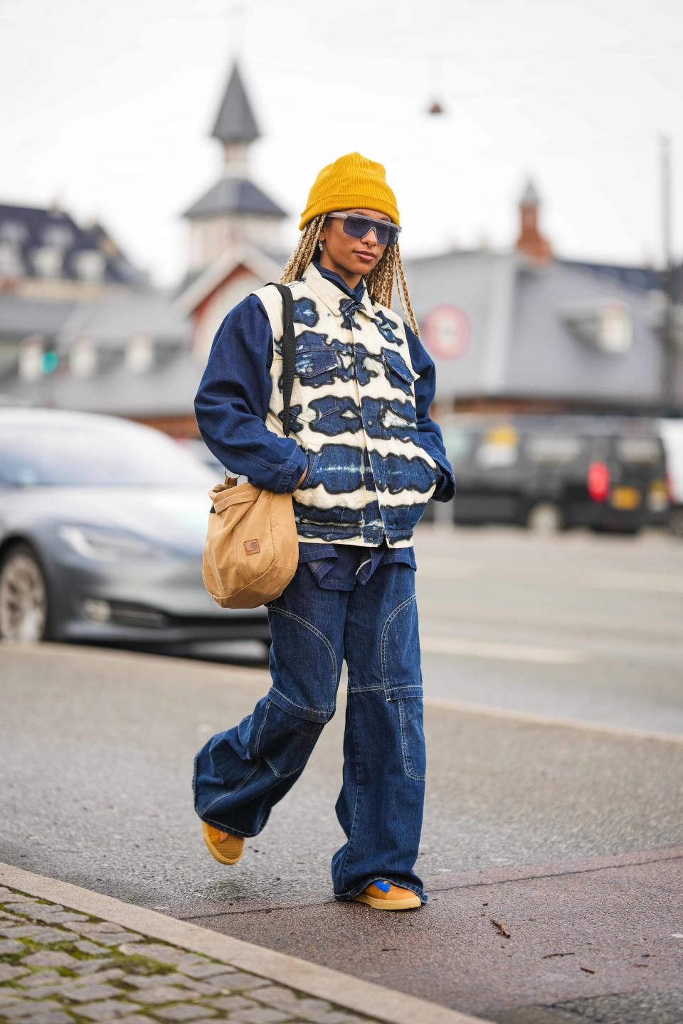 COPENHAGEN, DENMARK - FEBRUARY 01: A guest wears a yellow wool beanie, black futurist sunglasses, a navy blue shirt jacket, a white and navy blue tie and dye print pattern sleeveless denim jacket, navy blue embroidered seams pattern wide legs cargo pants, a brown denim shoulder bag from Carhartt, camel and blue print pattern fabric sneakers , outside OperaSport , during the Copenhagen Fashion Week Autumn/Winter 2023 on February 01, 2023 in Copenhagen, Denmark. (Photo by Edward Berthelot/Getty Images)