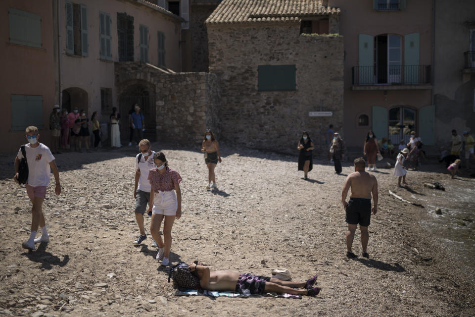 Tourists wearing masks walk by a sunbather in Saint-Tropez, southern France, Saturday Aug 8, 2020. The glamorous French Riviera resort of Saint-Tropez is requiring face masks outdoors starting Saturday, threatening to sober the mood in a place renowned for high-end, free-wheeling summer beach parties. (AP Photo/Daniel Cole)