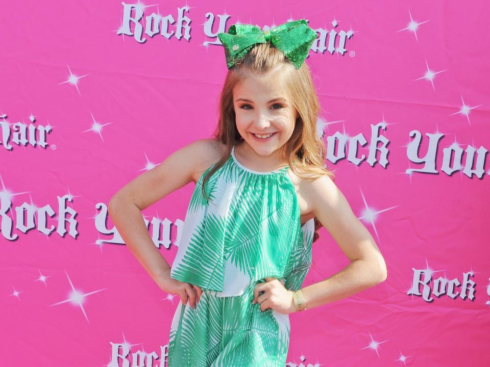 a young (approximately 10 years old) piper rockelle in a green shirt, shorts, and bow standing on a pink red carpet. wall behind her reads "rock your hair"