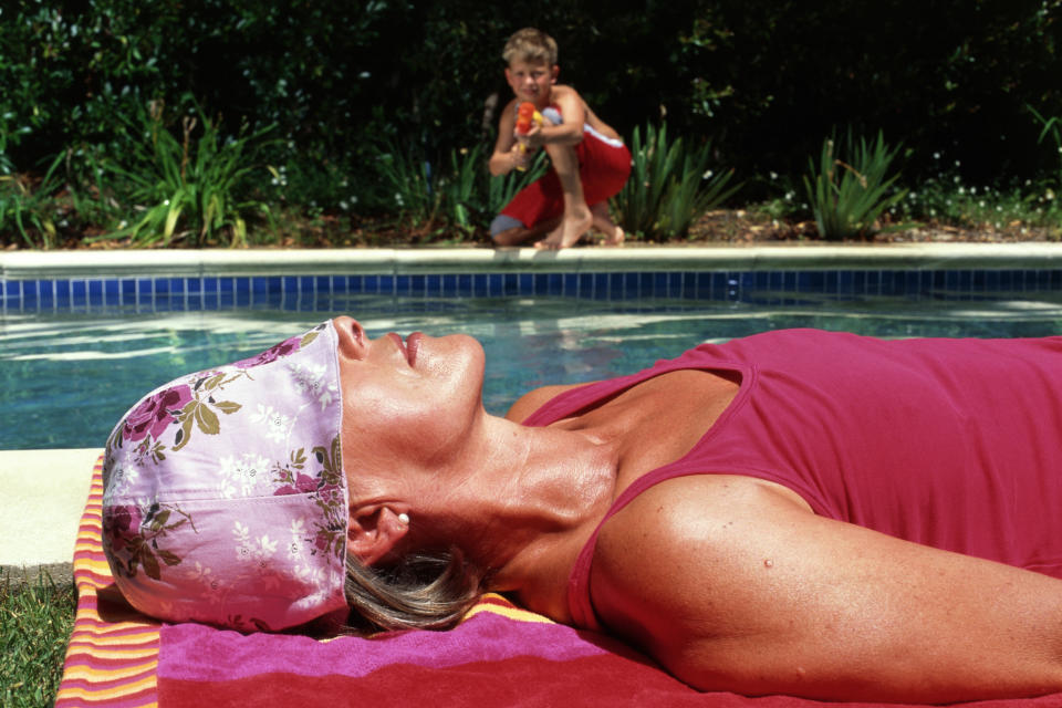 woman tanning by the pool by a kid across the pool aim a water gun at her