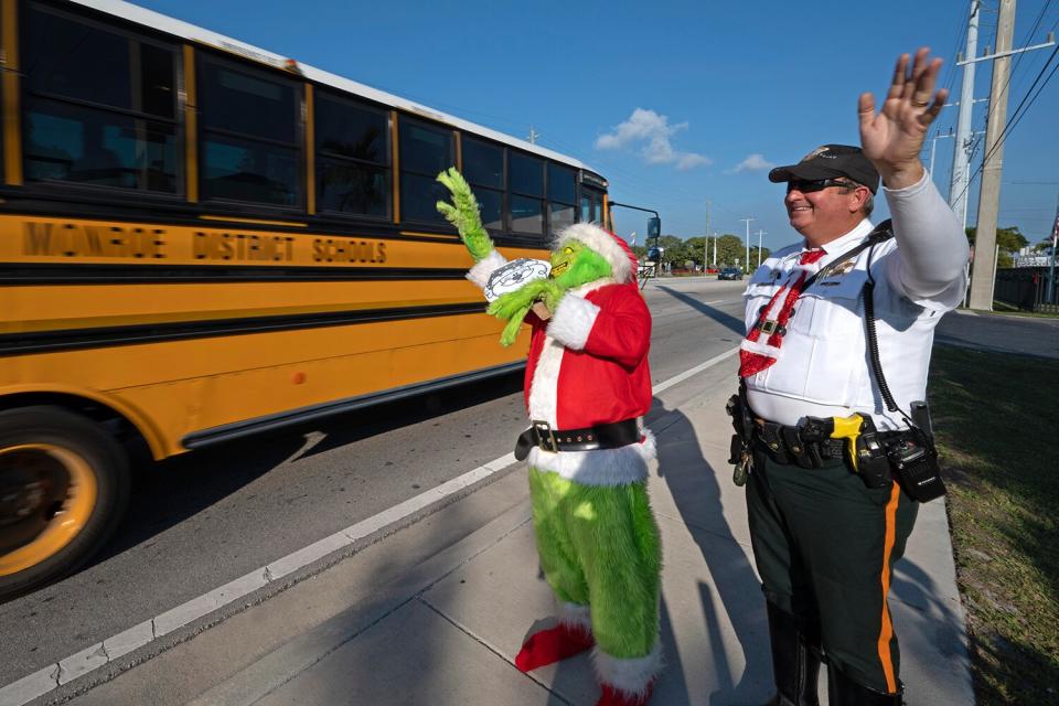 Lou Caputo, left, costumed as the Grinch, and Deputy Andrew Leird, right, wave at a school bus rolling on the Florida Keys Overseas Highway Tuesday, Dec. 13, 2022, in Marathon, Fla