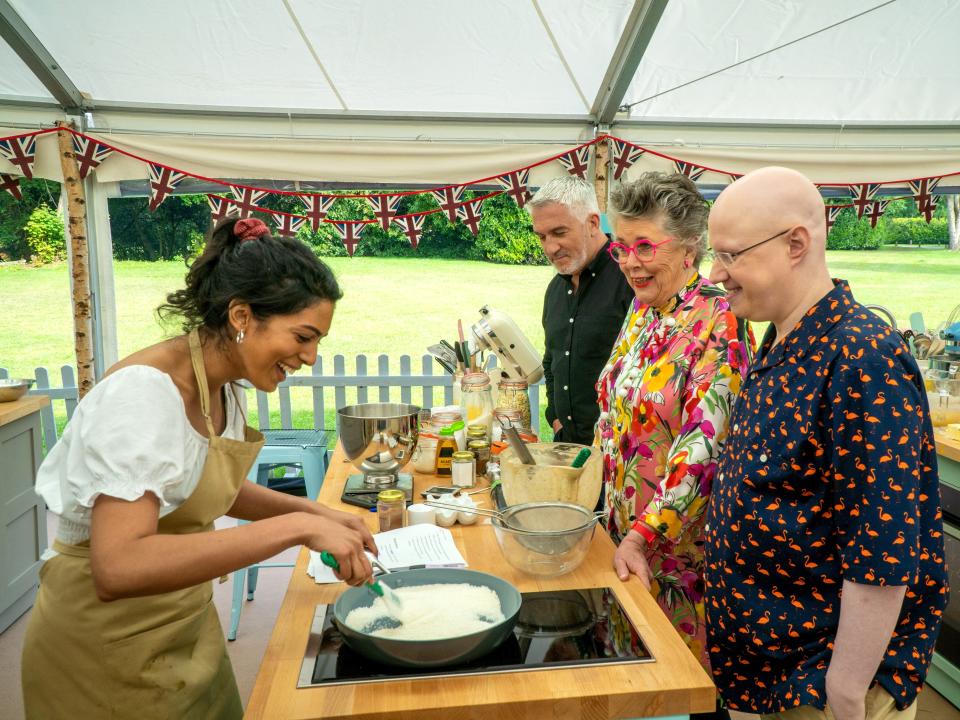 Crystelle baking while Paul, Prue, and Matt look over her and smile - Great British Baking Show / GBBO