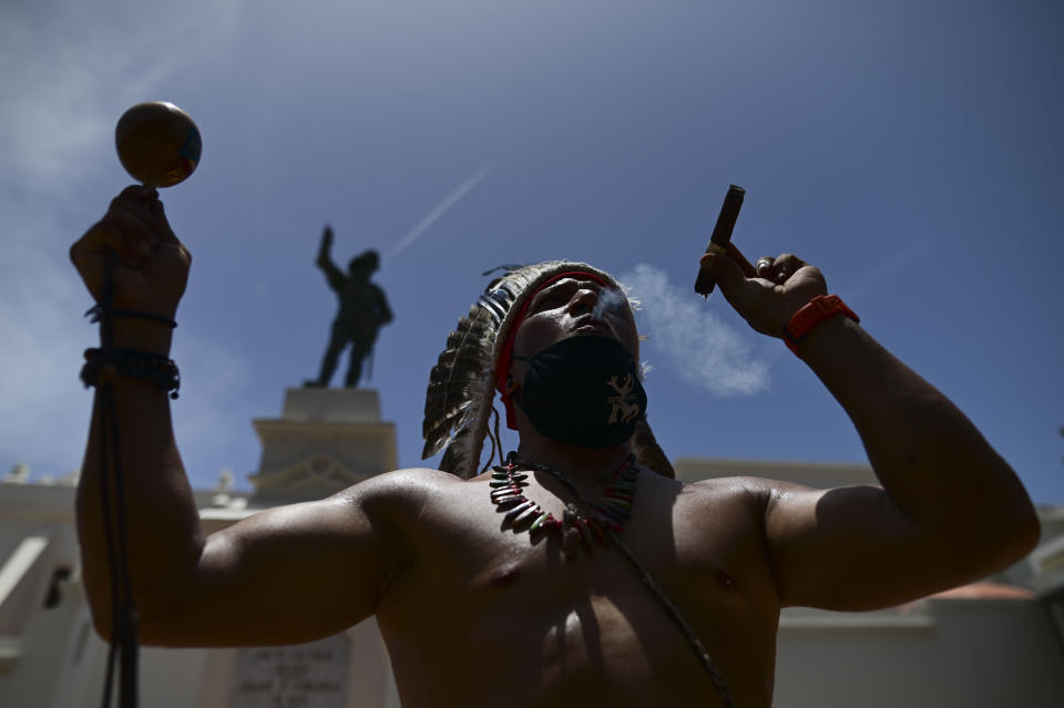 FILE - Gypsy Cordova blows smoke and shakes a maraca in front of a Juan Ponce de Leon monument while leading a group of activists in a march demanding statues and street names commemorating symbols of colonial oppression be removed, in San Juan, Puerto Rico, Saturday, July 11, 2020. Unknown people have toppled a statue of Spanish explorer Juan Ponce de León ahead of a visit of King Felipe VI to the U.S. territory of Puerto Rico. Col. José Juan García, police commissioner for San Juan, told The Associated Press on Monday, Jan. 24, 2022 that officers patrolling the cobblestone streets of the capital’s historic district hear a loud bang at 4:30 a.m. and found the statue broken in pieces. (AP Photo/Carlos Giusti, File)