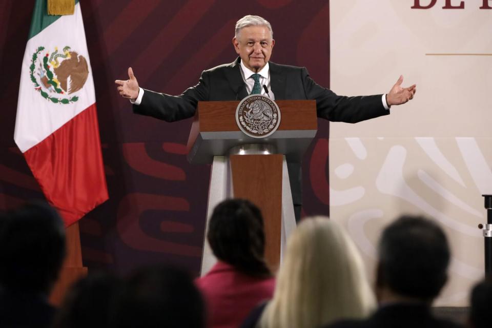 A man in a suit and tie stands at a lectern with his arms spread. A Mexican flag is displayed near him.