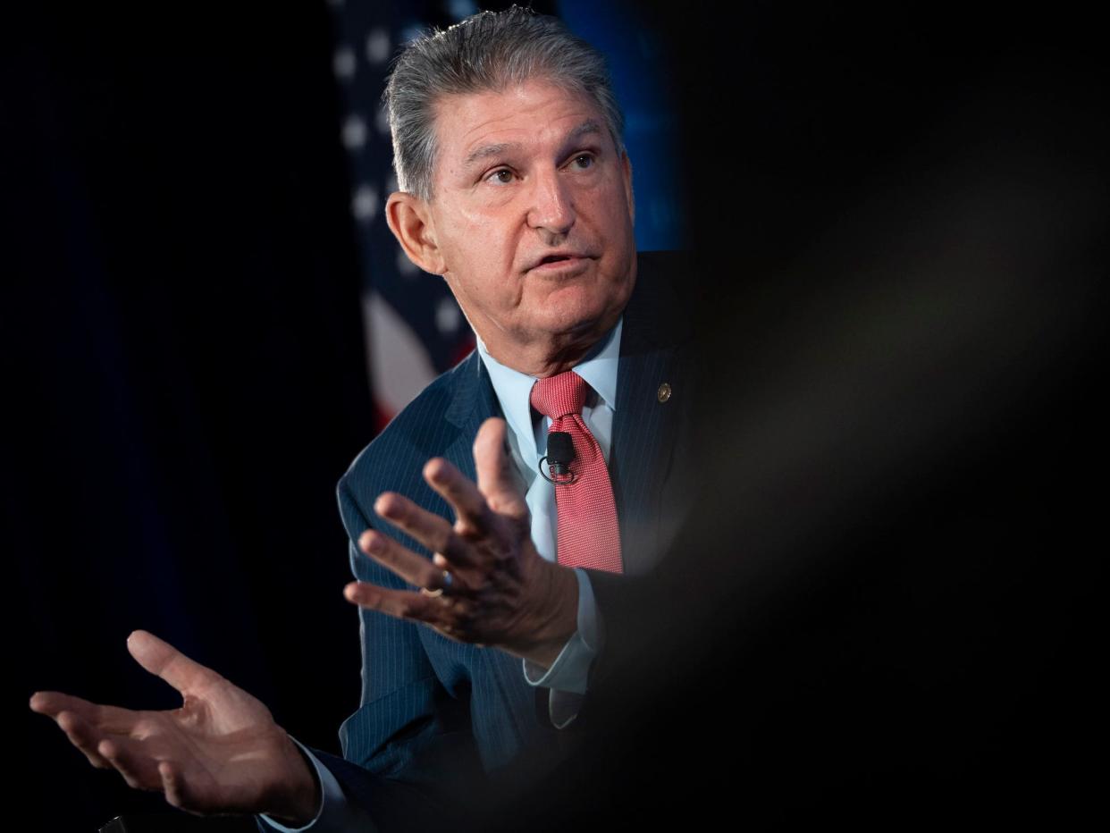 Senator Joe Manchin gestures in front of an American flag with his palms up, the right half of the photo is blurred out in black.