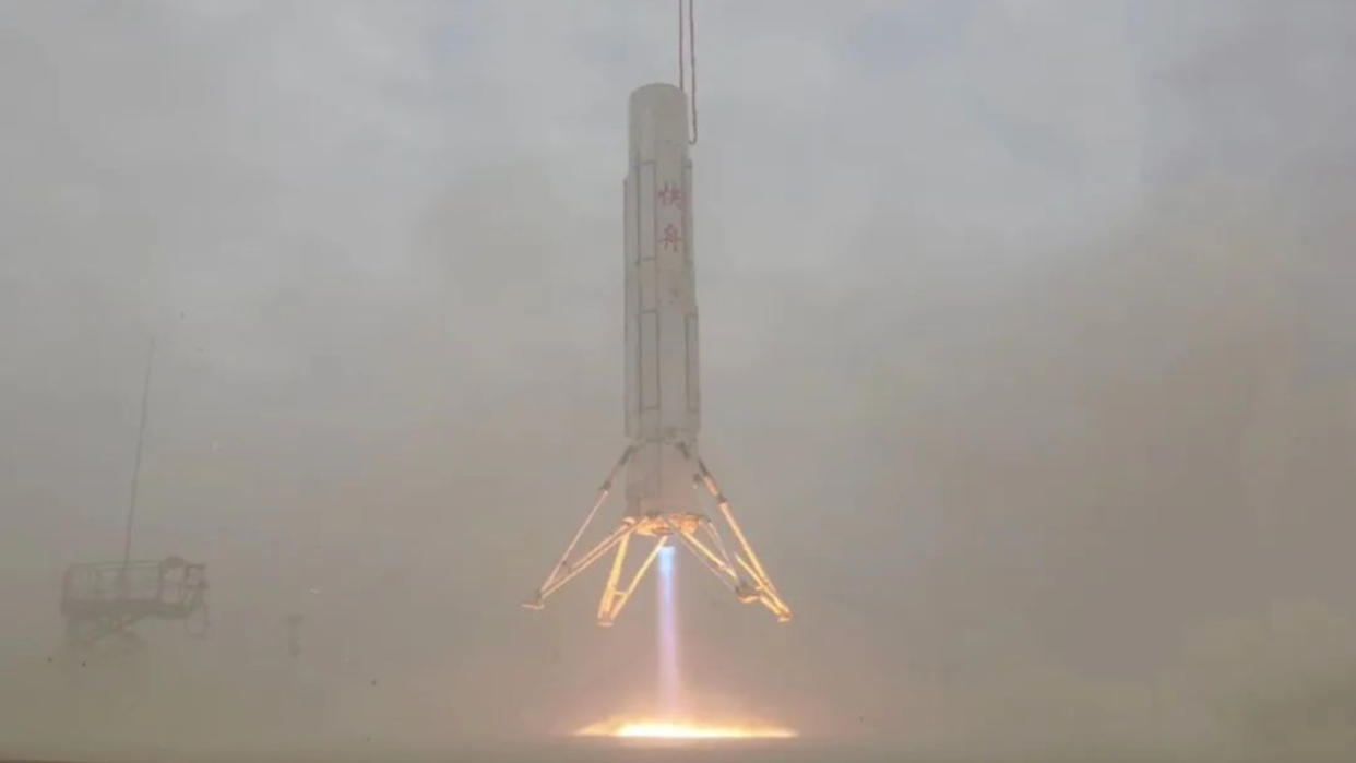  Close-up view of a white rocket hovering just above the ground, with blue flame blasting from its base. 