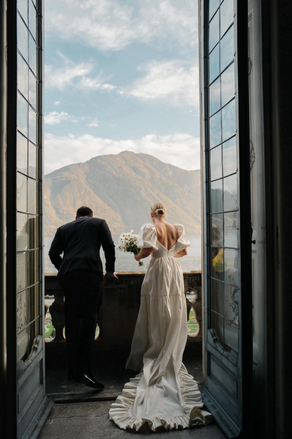 A bride and groom look down at a lake on a balcony in their wedding attire.