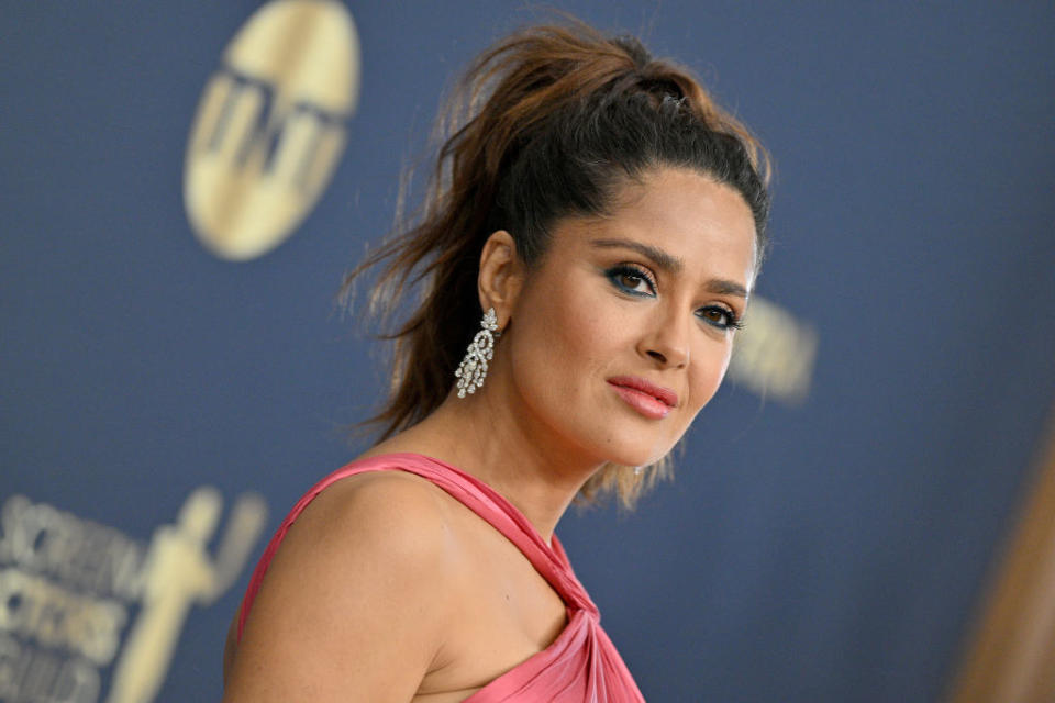 her hair is in a high ponytail, her eyes are highlighted with bold eyeliner in a contrasting color in her waterline, and silver chandelier earrings adorn her earlobes