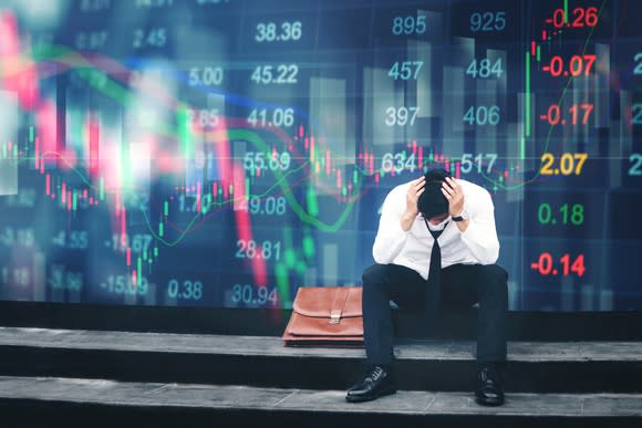 A man sits on a sidewalk with head in hands in front of a wall showing a declining share price chart.