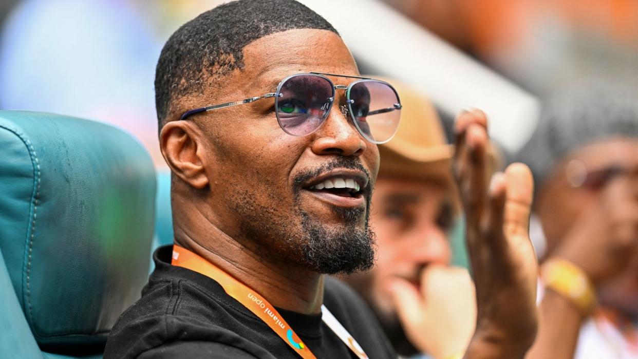 jamie foxx smiles and waves while seated, he wears sunglasses, a black t shirt and an orange lanyard