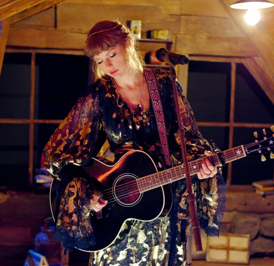 Taylor Swift in a patterned maxi dress playing acoustic guitar in a rustic room