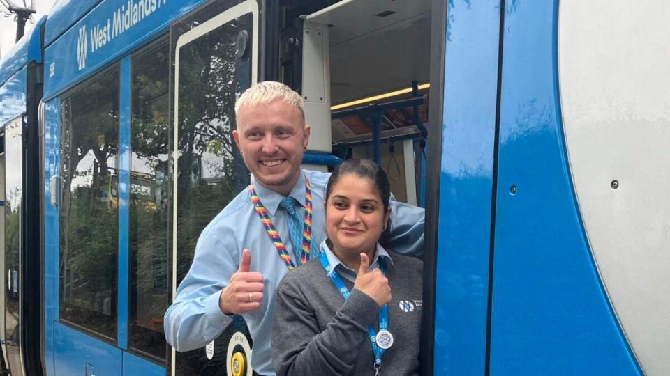 A man with bleached blond hair stands behind a woman with black hair. They are both hanging off the side of a blue tram as they smile and give thumbs up.