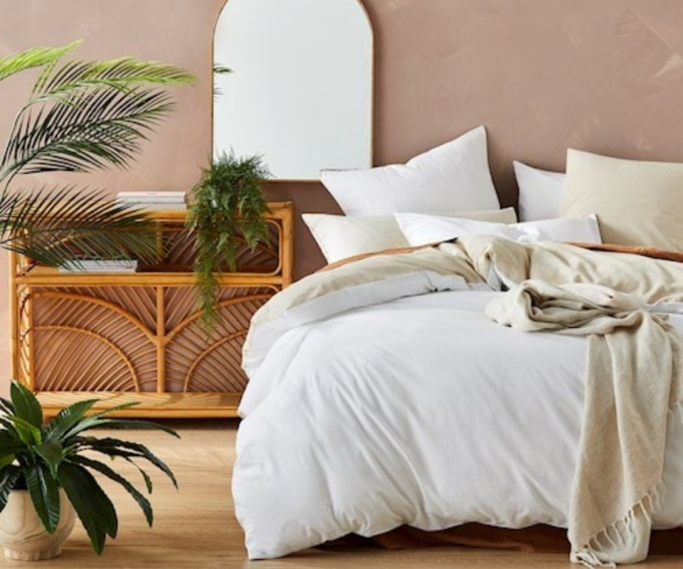 A bedroom with a mirror, indoor plants and a white bed behind a pale pink wall as a backdrop.