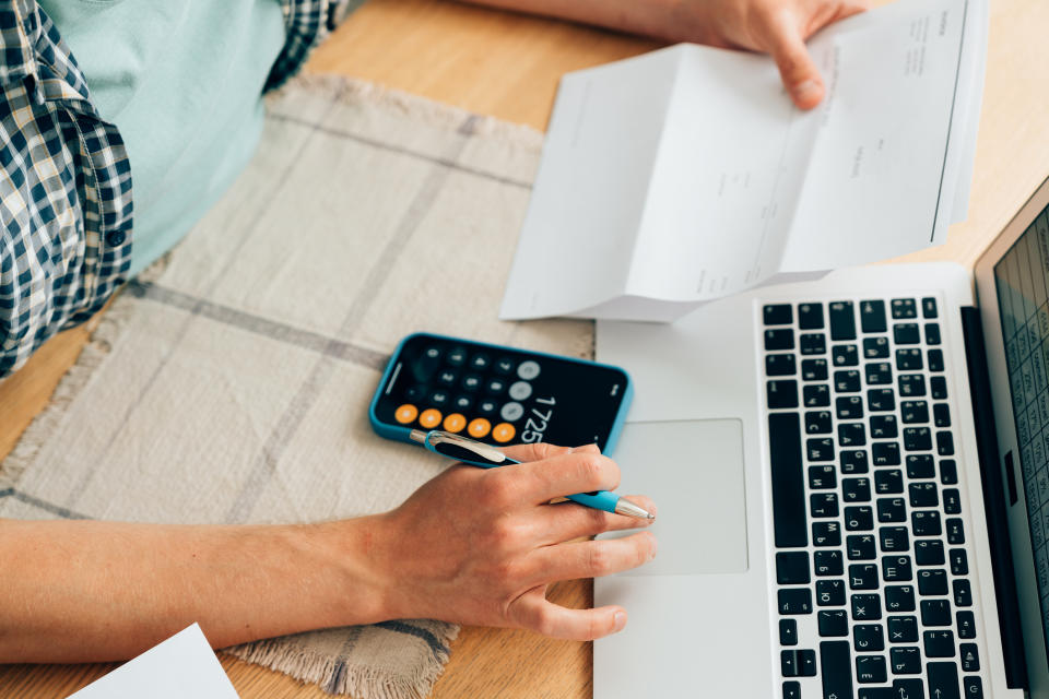 Person working at a desk with a laptop, holding a printed document, using a calculator app on their phone, and writing on a piece of paper