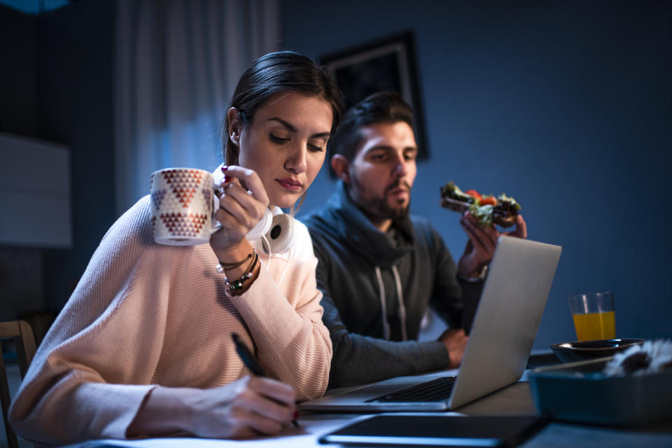 Young man and woman working from the comfort of their home at night. A woman is holding a cup of hot beverage while a man is eating a sandwich.