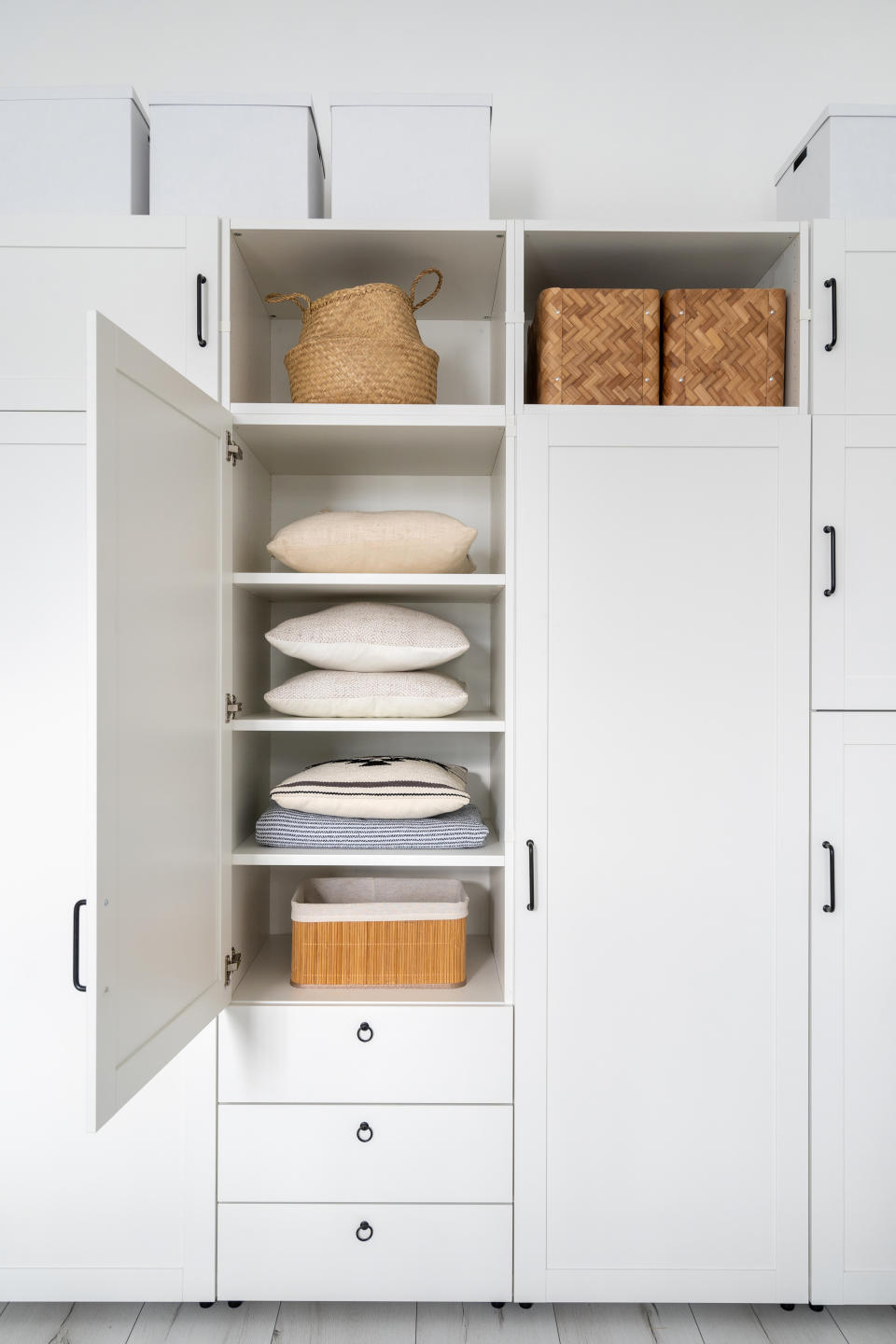 front view of modern white wardrobe with open door, pillows and blanket inside, wicker boxes on shelf in bedroom with light interior, organized storage concept