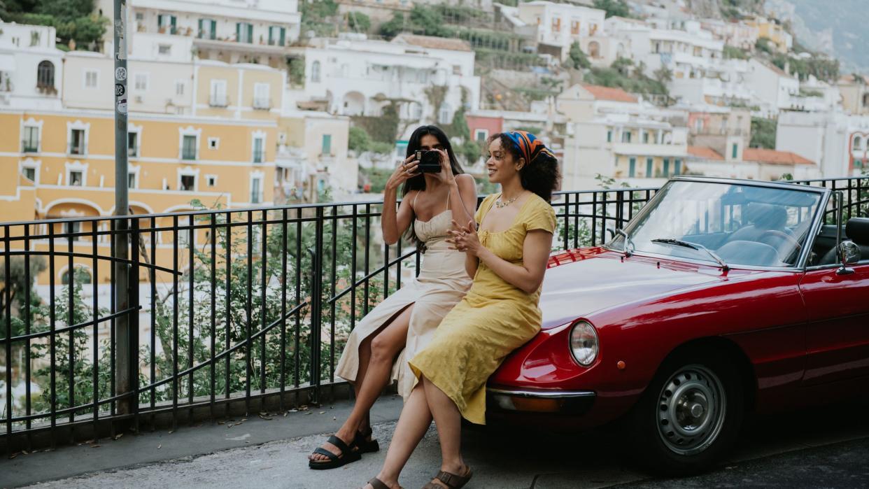 two woman sit on the bonnet of a vintage red convertible car enjoy touring italy together