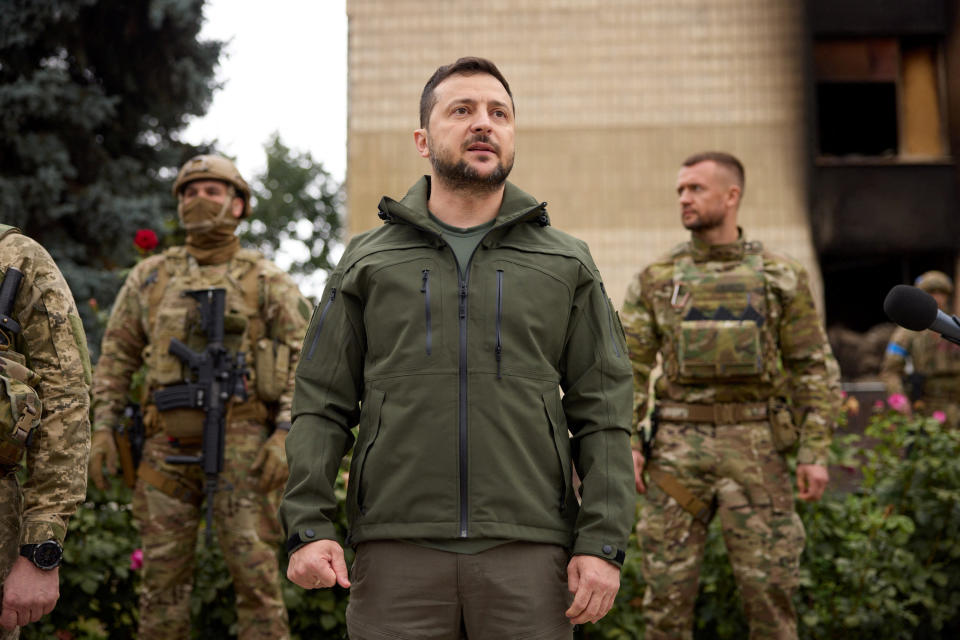 Volodymyr Zelensky stands near people wearing camouflage fatigues.