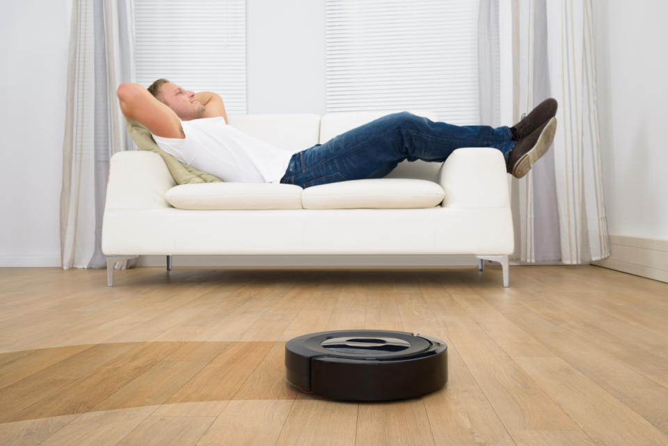 A man reclines on a couch while a robotic vacuum cleans the floor.